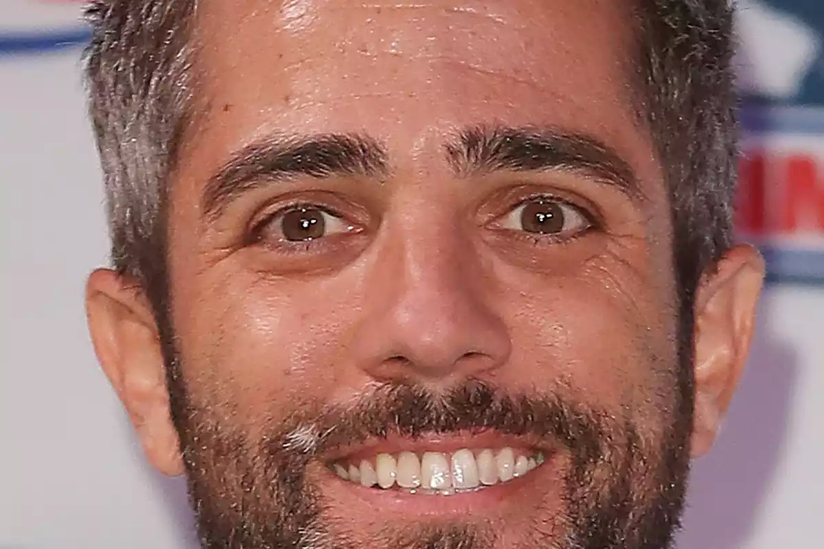 Roberto Leal con una sonrisa de compromiso, los dientes a la vista y un logo detrás